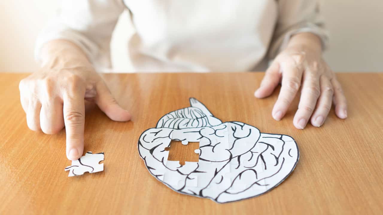 Eine ältere Frau sitzt vor einem Papierbild eines Gehirns und entfernt ein Puzzlestück